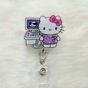 May include: A badge reel featuring a cartoon character with a pink bow, wearing a purple medical uniform, and holding a stethoscope. The character stands next to an ultrasound machine displaying an image. The reel has a clear plastic strap and a metal clip.