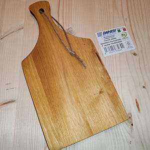 Può includere: Un tagliere in legno con manico e foro per appenderlo. La tavola è marrone chiaro con venature naturali. Un'etichetta con testo è attaccata. La tavola misura circa 40 cm di lunghezza.