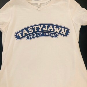 May include: White t-shirt with a crew neck and short sleeves. The shirt features a navy blue arched design with the words "TASTYJAWN" above "PHILLY FRESH" in white lettering. The shirt is made of a soft material.