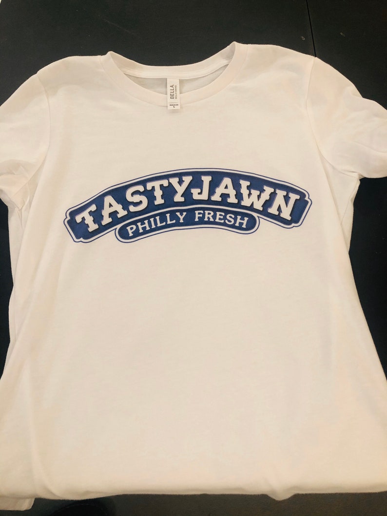 May include: White t-shirt with a crew neck and short sleeves. The shirt features a navy blue arched design with the words "TASTYJAWN" above "PHILLY FRESH" in white lettering. The shirt is made of a soft material.