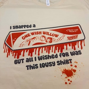 May include: A cream-colored t-shirt featuring a graphic of a red and white wishbone-shaped box labeled ONE WISH WILLOW with the text I snapped a and BUT all I WISHED FOR WAS This lousy SHIRT, accompanied by red paint-like drips and a splatter.