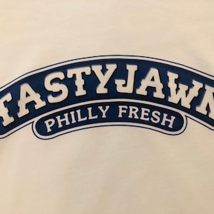May include: White t-shirt with a navy blue and white graphic. The graphic reads "TASTYJAWN" above "PHILLY FRESH" in a curved, arched design.