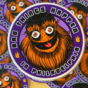 Könnte beinhalten: Ein runder Aufkleber mit lila Hintergrund und weißem Rand. Er zeigt ein Cartoon-Monster mit orangefarbenem Fell, einem schwarzen Hut und einem breiten Grinsen. Der Text auf dem Aufkleber lautet "Bad Things Happen in Philadelphia".
