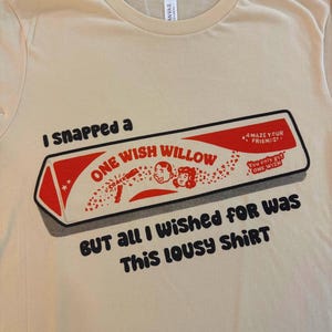 May include: Beige t-shirt with a graphic of a red and black candy wrapper that reads "ONE WISH WILLOW." Text on the shirt says "I snapped a...BUT all I wished for was This lousy shirt."