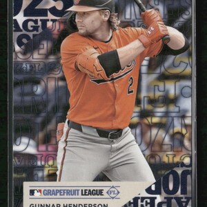May include: A baseball trading card featuring Gunnar Henderson of the Baltimore Orioles in an orange jersey, grey pants, and black helmet, holding a bat. The card includes the text Grapefruit League, Gunnar Henderson, and Baltimore Orioles.