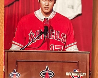 May include: A baseball card featuring Shohei Ohtani in a red Angels jersey, speaking at a podium. The card includes the Angels logo, the text "Opening Day," and the Topps RC logo. The jersey number 17 is visible.