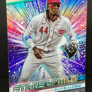May include: A baseball card featuring Elly De La Cruz of the Cincinnati Reds. The card has a holographic background with the text "STARS OF MLB". De La Cruz is wearing a white Reds uniform with the number 44.