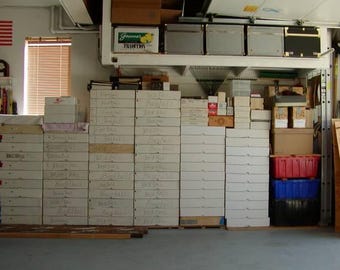 May include: A garage storage area with numerous white boxes stacked on the floor and shelves. The boxes are labeled with handwritten text. Red and blue storage bins are visible, along with an American flag and a window.