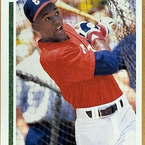 Puede incluir: Tarjeta de béisbol con Michael Jordan con camiseta roja y pantalón blanco, en pleno swing. Lleva una gorra azul con un logo blanco. La tarjeta tiene un borde verde y el texto "Upper Deck 91".