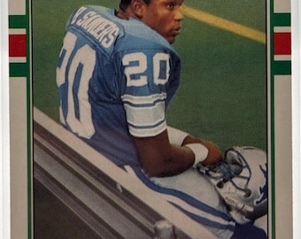 May include: A Topps trading card featuring Barry Sanders of the Detroit Lions. Sanders is wearing a blue and white jersey with the number 20. The card has a green border and the Topps logo.
