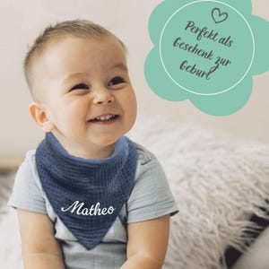Puede incluir: Un babero azul marino para bebé con el nombre "Matheo" en escritura blanca. El babero es triangular y está hecho de una tela texturizada. La imagen también incluye texto en forma de flor que dice "Perfekt als Geschenk zur Geburt".