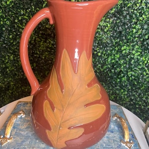 May include: A terracotta-colored ceramic pitcher with a large, embossed oak leaf design in shades of orange and brown. The pitcher has a curved handle and a spout for pouring. The pitcher is sitting on a blue and white tray with gold handles.