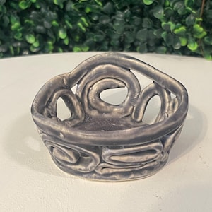 May include: A small, gray ceramic bowl with a unique, openwork design. The bowl features a series of swirling, raised patterns around the sides and top, creating an artistic and textured appearance. The bowl is sitting on a white surface.