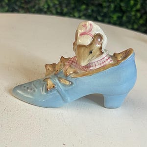 May include: A light blue ceramic shoe figurine with a mouse wearing a pink and white striped shirt and a white hat with a pink number 2. Several smaller mice are also in the shoe.