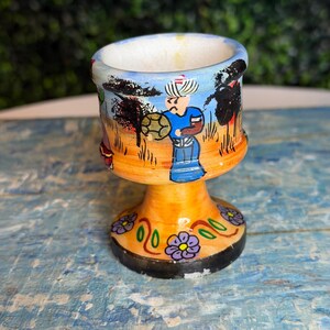 May include: A hand-painted ceramic egg cup with a vibrant design. The cup features a yellow base with floral accents and a detailed scene of figures in a landscape. The rim is blue, and the overall style is folk art.
