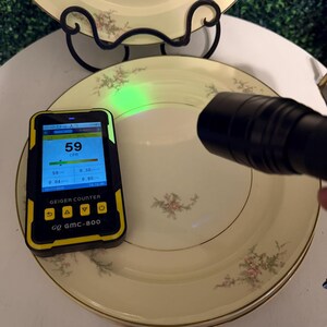 May include: A Geiger counter showing a reading of 59 CPM is placed near a stack of vintage plates. The plates are cream-coloured with a floral design and gold trim. A black torch is directed at the plates.