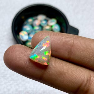 K&ouml;nnte beinhalten: Ein dreieckiger, schillernder Opal-Edelstein mit roten, gr&uuml;nen und gelben Blitzen. Der Opal wird zwischen Fingern gehalten, mit einem schwarzen Beh&auml;lter mit &auml;hnlichen Opalen im Hintergrund. Der Edelstein hat ein durchscheinendes, kristallines Aussehen.
