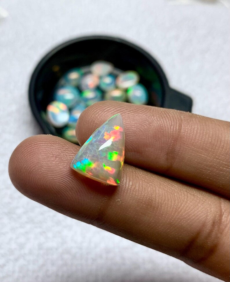 K&ouml;nnte beinhalten: Ein dreieckiger, schillernder Opal-Edelstein mit roten, gr&uuml;nen und gelben Blitzen. Der Opal wird zwischen Fingern gehalten, mit einem schwarzen Beh&auml;lter mit &auml;hnlichen Opalen im Hintergrund. Der Edelstein hat ein durchscheinendes, kristallines Aussehen.