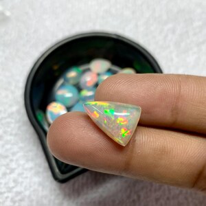 K&ouml;nnte beinhalten: Ein dreieckiger, schillernder Opal-Edelstein, der zwischen Fingern gehalten wird und gr&uuml;ne, orange und gelbe Blitze zeigt. Der Opal befindet sich vor einem verschwommenen Hintergrund aus anderen Opalen in einem schwarzen Beh&auml;lter.
