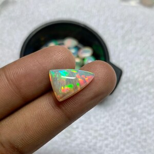 K&ouml;nnte beinhalten: Ein dreieckiger, schillernder Opal-Edelstein. Der Opal zeigt ein Farbspiel mit gr&uuml;nen, roten und gelben Blitzen. Der Edelstein wird zwischen Fingern gehalten, mit einem schwarzen Beh&auml;lter im Hintergrund.