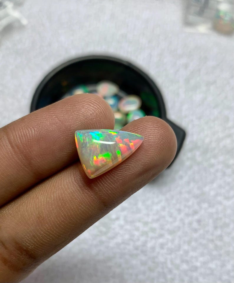 K&ouml;nnte beinhalten: Ein dreieckiger, schillernder Opal-Edelstein. Der Opal zeigt ein Farbspiel mit gr&uuml;nen, roten und gelben Blitzen. Der Edelstein wird zwischen Fingern gehalten, mit einem schwarzen Beh&auml;lter im Hintergrund.