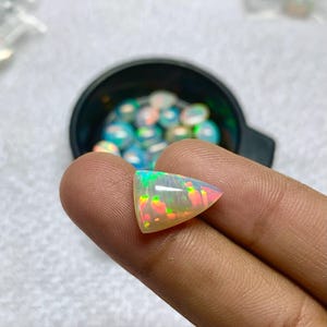 K&ouml;nnte beinhalten: Ein dreieckiger, schillernder Opal-Edelstein mit gr&uuml;nen, rosa und gelben Lichtreflexen. Der polierte Stein wird zwischen Fingern gehalten, mit einer schwarzen Schale mit &auml;hnlichen Opalen im Hintergrund. Die Oberfl&auml;che des Opals reflektiert das Licht.