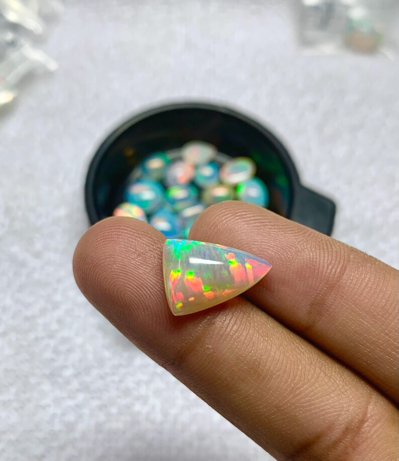 K&ouml;nnte beinhalten: Ein dreieckiger, schillernder Opal-Edelstein mit gr&uuml;nen, rosa und gelben Lichtreflexen. Der polierte Stein wird zwischen Fingern gehalten, mit einer schwarzen Schale mit &auml;hnlichen Opalen im Hintergrund. Die Oberfl&auml;che des Opals reflektiert das Licht.