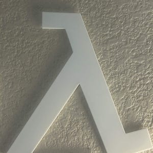 May include: A white, three-dimensional wall-mounted sign featuring the Greek letter lambda, which serves as the logo for the video game series Half-Life. Below the large lambda symbol is a rectangular plaque with the text Half-Life embossed on it.