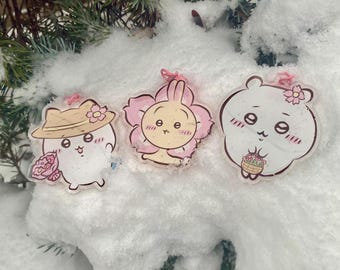 Puede incluir: Tres colgantes de acrílico transparente con personajes de dibujos animados. Uno es un oso blanco con flores, otro un conejito en una flor y el tercero un personaje con sombrero y flores. Cada colgante tiene una cinta rosa.