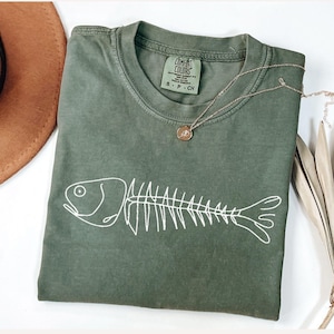 以下が含まれることがあります： 白い魚の骨格グラフィックが特徴のオリーブグリーンのTシャツ。 シャツはクルーネックと半袖です。 ペンダント付きのゴールドネックレスがシャツの上にあります。 茶色の帽子が左上にあります。