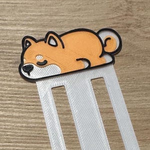 Könnte beinhalten: Weißer Lesezeichen mit einem schlafenden Shiba Inu-Hund-Design. Der Hund ist orange und weiß mit schwarzen Akzenten. Das Lesezeichen besteht aus einem weißen, strukturierten Material.