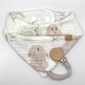May include: A white and beige baby bib with a bunny print and a pacifier. The bib has a snap closure and a wooden teether with a fabric loop. The bib is made of soft, textured fabric.