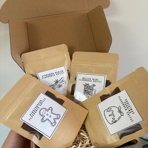 May include: A brown cardboard box filled with four kraft paper pouches of flavored coffee. Each pouch has a white label with a black illustration and text. Flavors include Cinnamon Danish, Mulled Wine, Gingerbread, and Christmas Pudding.