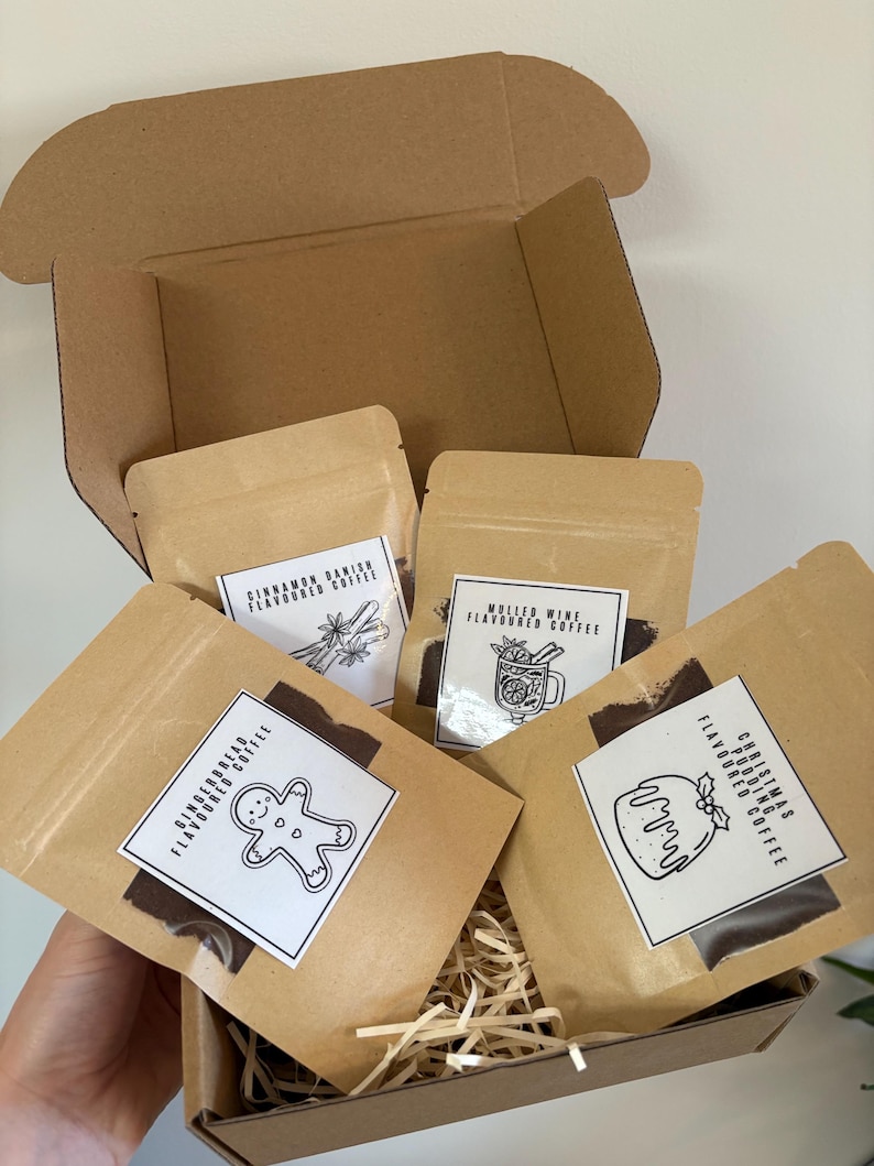 May include: A brown cardboard box filled with four kraft paper pouches of flavored coffee. Each pouch has a white label with a black illustration and text. Flavors include Cinnamon Danish, Mulled Wine, Gingerbread, and Christmas Pudding.