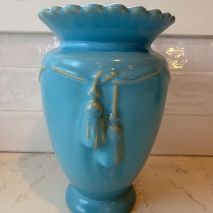 May include: A bright turquoise ceramic vase featuring a scalloped rim and a raised relief design of draped rope with two hanging tassels. The glaze has a smooth, glossy finish with subtle yellow-toned highlights along the embossed details.