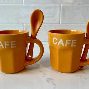 May include: Two orange ceramic mugs with the word "CAFE" in white lettering. Each mug has a matching orange spoon resting on the handle. The mugs have a geometric design and are suitable for coffee or tea.