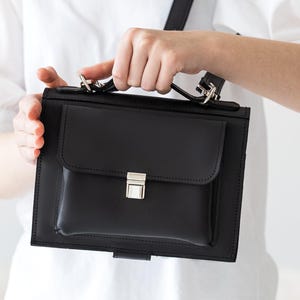 May include: Black leather crossbody bag with a rectangular shape and a front flap pocket. The bag features a silver-colored clasp and a long, adjustable strap. The bag is held by a person wearing a white shirt.