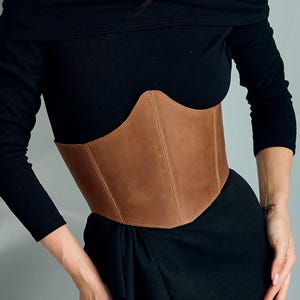 May include: A tan leather corset belt worn over a black off-the-shoulder long-sleeved top and a matching black skirt. The corset features a structured, pointed front panel with visible vertical stitching and a smooth, matte finish.