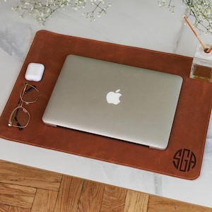 May include: A brown leather desk mat with a laptop, glasses, and wireless earbuds. The mat has a monogram in the lower right corner. A diffuser and flowers are in the background.