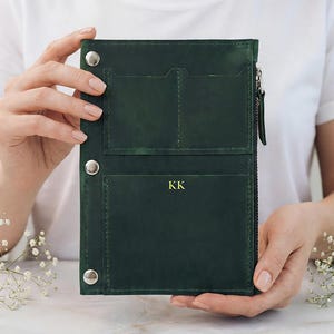 Pode incluir: Diário de couro verde escuro com fecho de correr e fechos de pressão. O diário tem dois bolsos interiores e um bolso frontal com as iniciais "KK" em letras douradas. O diário é mantido aberto por uma pessoa.