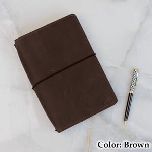 May include: A brown leather journal with a dark elastic closure and the initials "AG" embossed on the cover. A black and gold pen rests beside the journal. The journal is on a white marble surface.