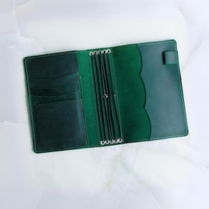 May include: Open, flat lay of a dark green leather journal. The journal is open, revealing multiple card slots on the left and a scalloped edge on the right. The journal has a snap closure and metal rings for holding inserts.