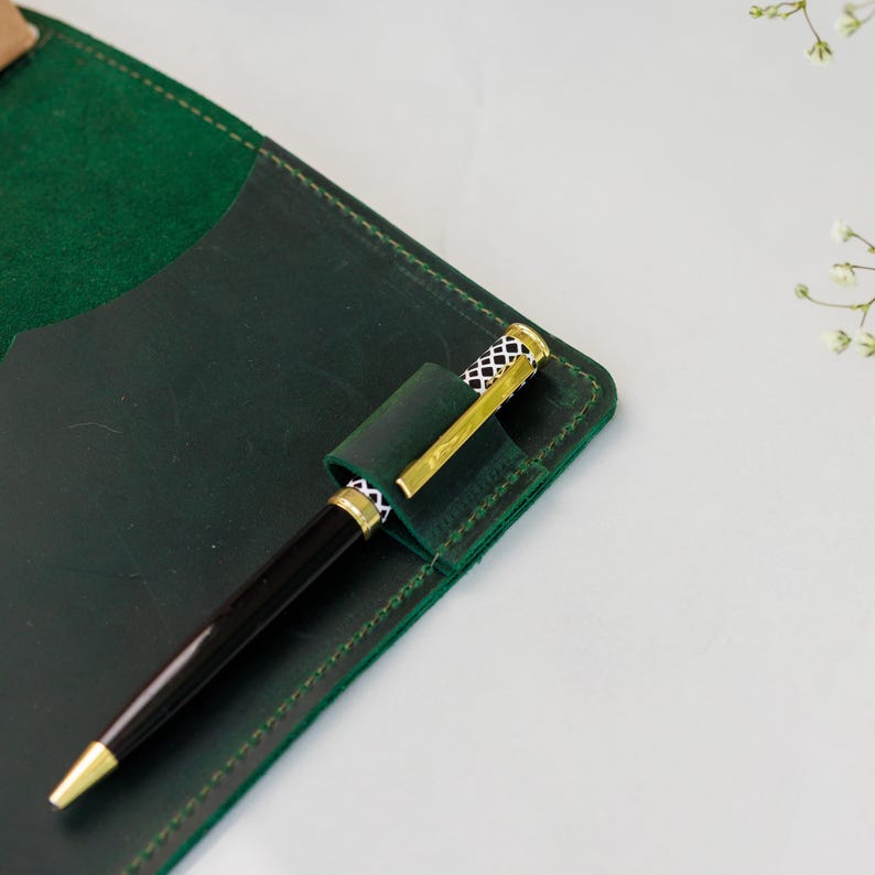 Pu&ograve; includere: Una penna nera con dettagli dorati &egrave; inserita in un portapenne in pelle verde scuro attaccato a un taccuino abbinato. Il portapenne presenta un motivo decorativo. Il taccuino &egrave; aperto, rivelando una tasca.