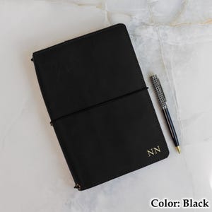 May include: A black leather journal with an elastic closure and gold initials "NN". A silver and black patterned pen rests beside the journal. The journal is open, revealing the interior pages. Color: Black.