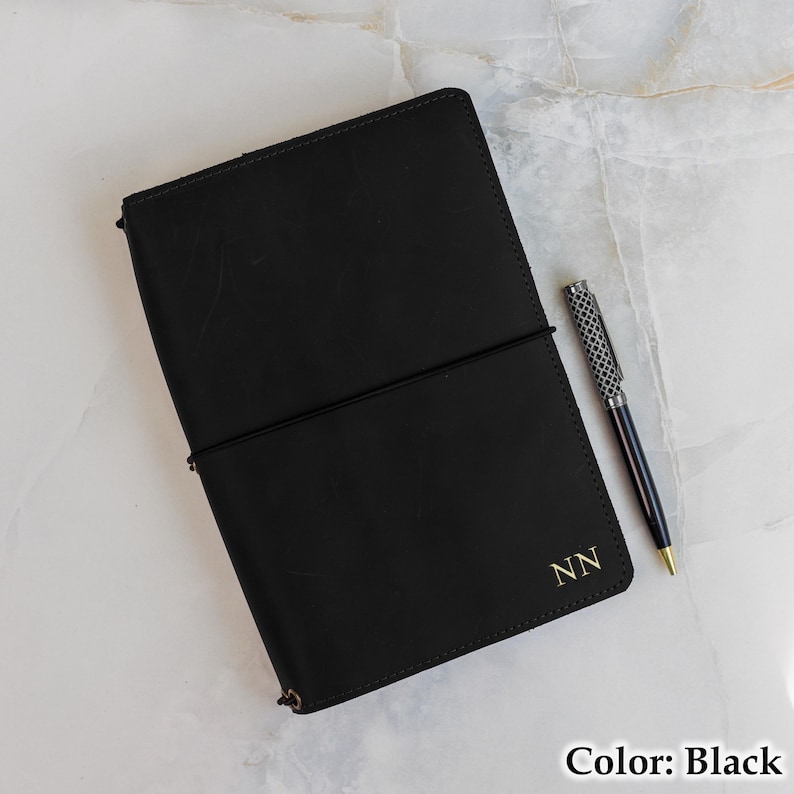May include: A black leather journal with an elastic closure and gold initials "NN". A silver and black patterned pen rests beside the journal. The journal is open, revealing the interior pages. Color: Black.