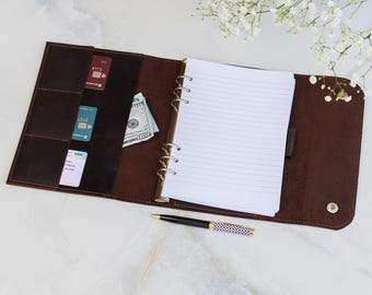 May include: Open brown leather binder with a lined notepad, credit cards, and cash. A black and white patterned pen rests beside the open binder. The binder has a snap closure and multiple pockets for organization.
