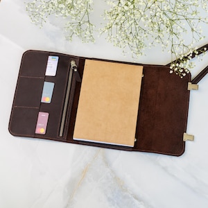 Peut inclure: Journal en cuir marron foncé ouvert, avec une fermeture éclair et une sangle. Le journal contient un carnet marron clair et des fentes pour cartes. Le journal est sur une surface en marbre blanc avec des fleurs blanches.