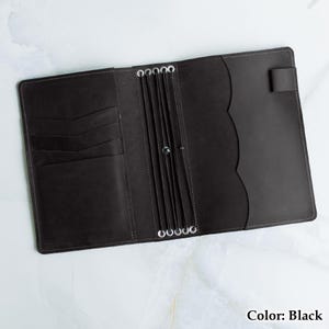 Pu&ograve; includere: Diario in pelle nera aperto con pi&ugrave; fessure per carte e un bordo smerlato. Il diario ha anelli argentati per tenere le pagine e una chiusura con cinturino. Il testo "Color: Black" &egrave; visibile.
