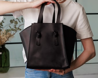 Puede incluir: Un bolso tote de cuero negro con dos asas superiores y nudos decorativos. El bolso tiene un diseño estructurado y lo sostiene una persona que lleva una camiseta blanca y vaqueros azules. Un accesorio elegante.