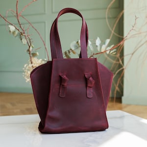 May include: A burgundy leather tote bag with two top handles and decorative knots. The bag has a structured design with a rectangular base and flared sides. The bag is made of a smooth, matte leather.